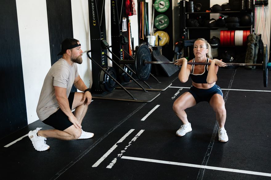 A coach kneeling beside an athlete mid-squat, watching her position